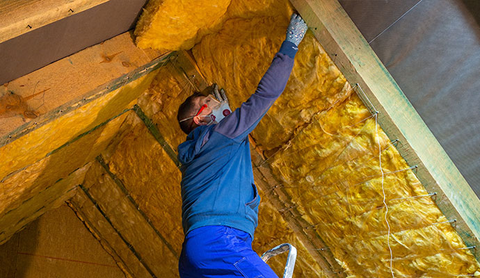 Technician in a respirator installing fiberglass insulation between roof rafters